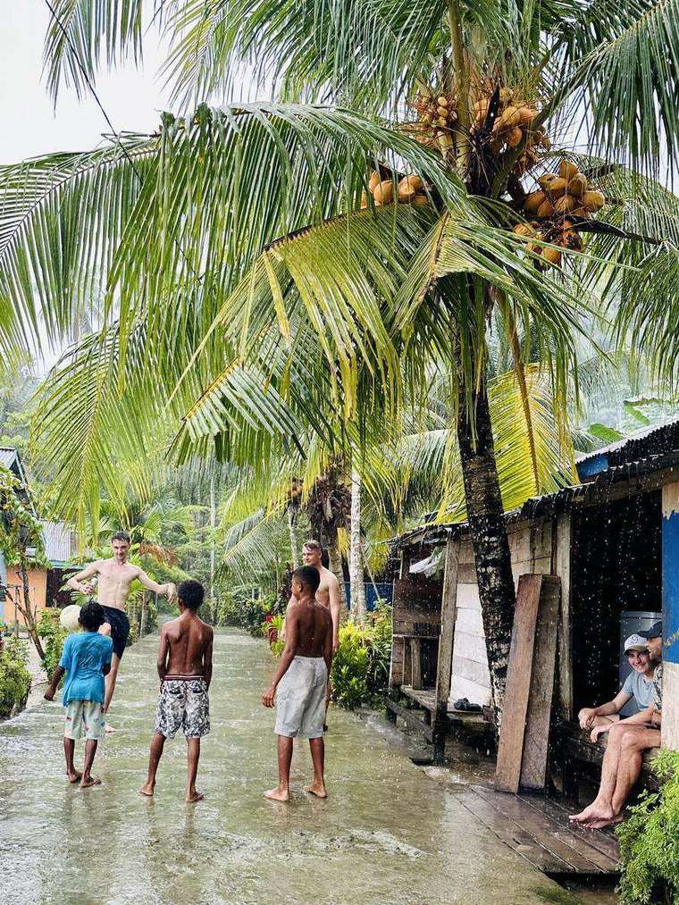 Kinder spielen mit jungen Besuchern Fußball auf einer nassen Dorfstraße unter Kokospalmen in Raja Ampat